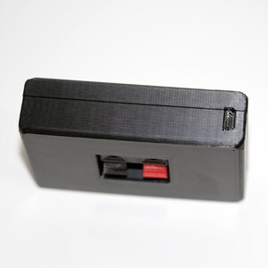 May include: A black rectangular electronic component with a textured surface. It features a red and black port and a small latch on the side. The component is likely used in audio or electronic devices.