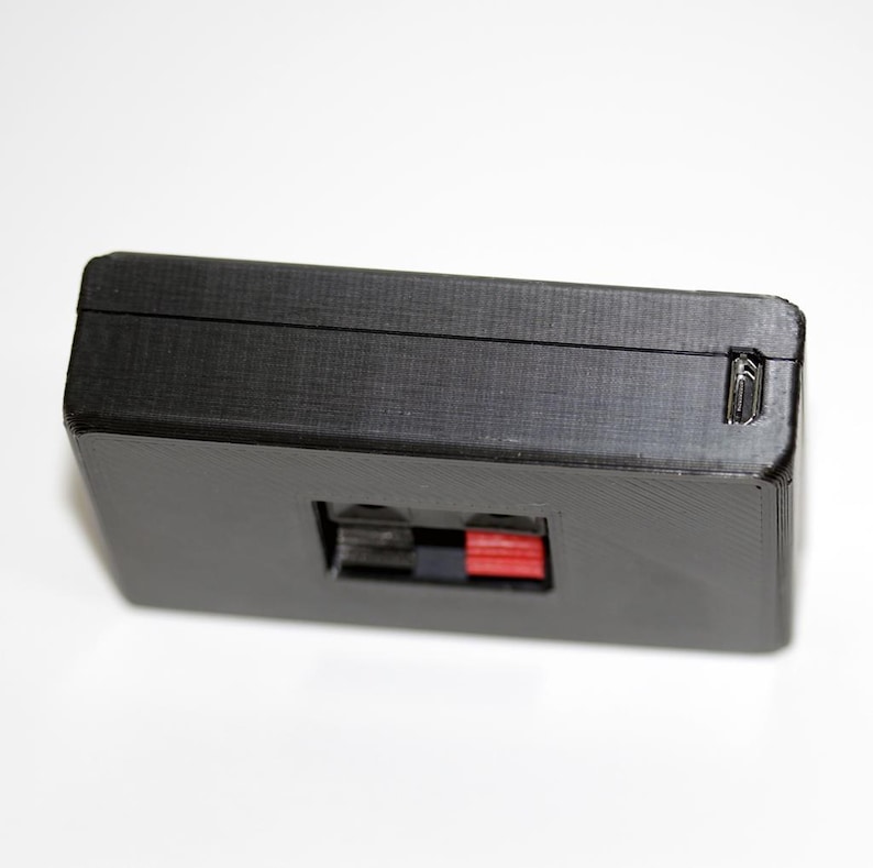 May include: A black rectangular electronic component with a textured surface. It features a red and black port and a small latch on the side. The component is likely used in audio or electronic devices.