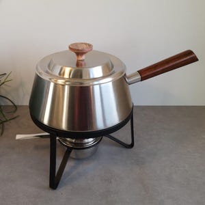 Può includere: Una pentola per fonduta in acciaio inossidabile con manico e coperchio in legno. La pentola poggia su un supporto in metallo nero con un bruciatore di combustibile sottostante. Il coperchio ha un pomello in legno. Il manico è marrone scuro.