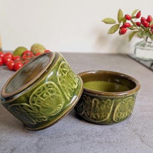 May include: Two round ceramic ramekins featuring a glossy olive green glaze and a raised, embossed pattern of repeating circular motifs. The interior of each bowl is a lighter, mottled lime green, while the exterior rims are a dark brown.