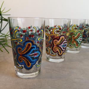 Op de afbeelding: Een set heldere glazen met een levendig, meerkleurig paisley-patroon. Het ontwerp bevat ingewikkelde krullen in blauwe, gele, rode en groene tinten, met kleine bloemaccenten die de bovenrand van elk glas sieren.
