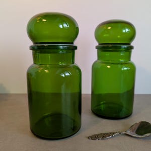 Op de afbeelding: Twee bijpassende cilindrische glazen potten in een levendige smaragdgroene kleur, elk voorzien van een afgerond, koepelvormig deksel. Een kleine, versierde zilverkleurige metalen lepel met gedetailleerd reliëf ligt naast de potten.