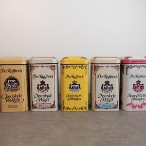 Peut inclure: Cinq boîtes en étain vintage De Ruijter's avec différents motifs et saveurs de chocolat. Les boîtes sont jaunes, blanches et roses avec des accents dorés. Le texte sur les boîtes indique "De Ruijter's" et comprend les saveurs "Chocolade Mokken Melk", "Chocolade Hagel Puur", "Gestampte Muisjes", "Chocolade Hagel Melk" et "Rose & Witte Muisjes".