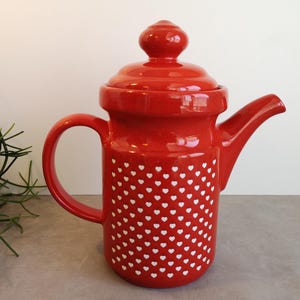 Op de afbeelding: Een felrode keramische theepot met een herhalend patroon van kleine witte hartjes op het cilindrische lichaam. De theepot heeft een bijpassend rood deksel met een ronde knop, een gebogen tuit en een stevig handvat aan de zijkant.