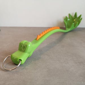 Op de afbeelding: Een groene plastic spaghettilepel in de vorm van een draak. De lepel heeft oranje stekels langs de rug en een zilveren metalen ring aan het uiteinde van de handgreep. De lepel ligt op een grijze ondergrond.