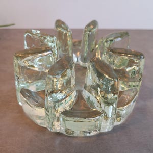 Op de afbeelding: Een kandelaar van helder glas met een ronde voet en acht dikke, rechtopstaande, getextureerde glazen segmenten in een ringvorm. Het glas heeft een licht groenachtige tint en een zwaar, solide uiterlijk met onregelmatige vlakken.