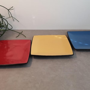 Op de afbeelding: Drie vierkante keramische borden met zwarte randen. De borden zijn rood, geel en blauw.