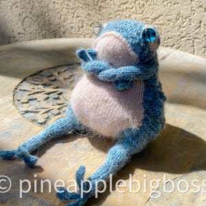 May include: A blue and pink knitted frog toy with large blue eyes. The frog is sitting on a wooden table with a carved design.