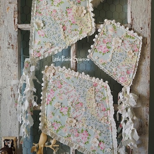 May include: Decorative fabric pieces with floral patterns in pink, blue, and white, adorned with lace trim and floral embellishments. The pieces are hanging on a rustic, white-painted wooden frame with chicken wire backing. The text "Little Prairie Sparrow" is visible.