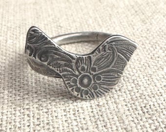 Puede incluir: Un anillo plateado con un diseño floral. El anillo presenta un diseño abierto con una flor central y patrones de hojas detallados. El anillo está sobre un fondo neutro y texturizado.