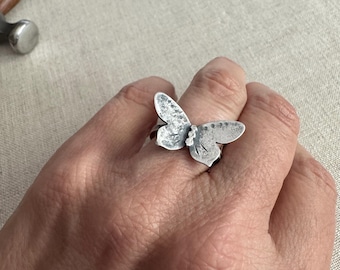 Könnte beinhalten: Ein silberner Schmetterlingsring. Der Ring hat ein detailliertes Schmetterlingsdesign mit strukturierten Flügeln und einem zarten Körper. Der Ring wird an einem Finger getragen und zeigt die filigrane Handwerkskunst des Schmucks.