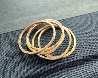 May include: Four gold-colored, thin, oval rings are intertwined on a dark gray textured surface. The rings have a slightly hammered texture, catching the light and creating highlights. The rings are likely jewelry.