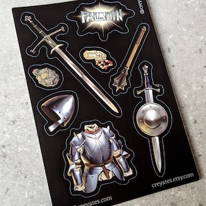 以下が含まれることがあります： ファンタジーをテーマにしたイラストが描かれた黒いステッカーシート。長剣、メイス、剣付きの盾、ヘルメット、鎧、手榴弾、ビーズが含まれています。「PALADIN」の文字が上部にあります。