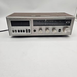 May include: A vintage AM/FM stereo electronic clock radio with a silver face and a wood-grain top. The radio features a digital display, various control knobs, and the brand name "SOUNDESIGN". The radio has a power cord.