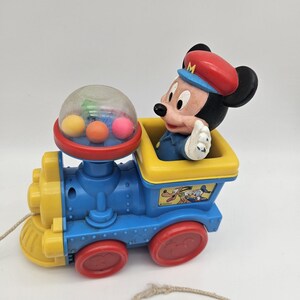 May include: A vintage pull-along toy train featuring Mickey Mouse as the conductor. The train is primarily blue and yellow with red wheels. The train car has a clear dome with colorful balls inside.