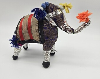 May include: A decorative elephant figurine with a purple and gold patterned body, adorned with silver accents and colorful tassels. The elephant has a striped saddle and is holding an orange pom-pom in its trunk. The figurine is approximately 6 inches tall.