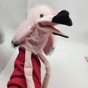 May include: A pink flamingo puppet with a black beak and red accents. The puppet has a red body and long, pink legs. The flamingo's head is made of soft, plush material.
