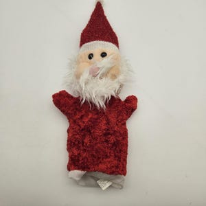 May include: A plush Santa Claus hand puppet. The puppet features a red textured body and hat with a white trim and pom-pom. The face is beige with black eyes, a pink nose, and a white beard. The puppet's arms are outstretched.