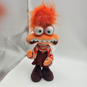 May include: A plush doll featuring bright orange hair, large protruding eyes with green pupils, and an orange face with a wide, surprised expression. The doll wears a striped orange and white long-sleeve shirt, dark brown overalls, and dark brown shoes.