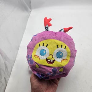 May include: A round plush toy featuring a yellow character face with large blue eyes, a wide smile, and freckles, encased in a pink hood decorated with purple flowers. Two red and white striped antennae-like appendages extend from the top.