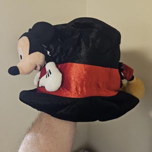 May include: A plush Mickey Mouse hat with a black top and a red band. The hat features a stuffed Mickey Mouse figure with a beige face, black ears, and white gloves. The hat is a fun accessory.