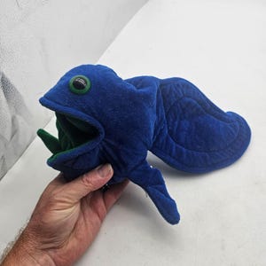 May include: A blue plush fish-shaped oven mitt with a wide open mouth, green interior lining, and a large green eye. The exterior features a soft, textured fabric with a rounded tail fin and a side fin, designed to function as a kitchen accessory.