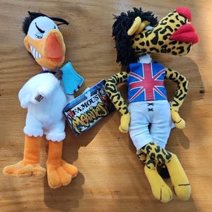 May include: Two plush toys: a white duck with orange feet and beak, and a leopard-print figure wearing a Union Jack vest and white pants. The duck has a tag that reads "Famous Monies". The leopard figure has black hair and red lips.