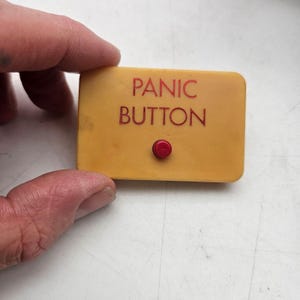 May include: A rectangular yellow plastic device with rounded corners featuring the text PANIC BUTTON printed in red letters. A small, circular red button is positioned directly below the text on the front face of the device.