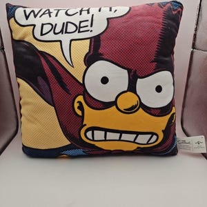 May include: Square decorative pillow featuring a comic book style image of a cartoon character with yellow skin, a red outfit, and the text "WATCH IT, DUDE!". The pillow has a red and yellow background.