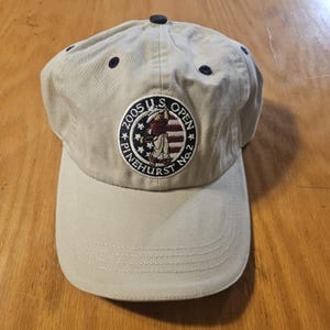 May include: Tan baseball cap with a curved brim and a button on top. The cap features a circular patch with the text "2005 U.S. OPEN PINEHURST No.2" and an illustration of a golfer.