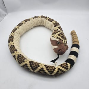 May include: A plush rattlesnake toy featuring a tan and brown diamond-patterned body, a dark brown head with black eyes, a black felt tongue, and a segmented tail with alternating black and tan bands, curled into a circular shape on a white surface.