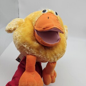 May include: A plush yellow duck puppet with an orange beak, feet, and legs. The puppet has black eyes and a pink interior mouth. The puppet is made of soft, fuzzy material and is designed to be worn on the hand.