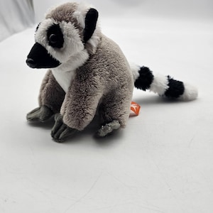 May include: A plush lemur toy with gray, white, and black fur. The lemur has a black nose, black eyes, and a striped tail. The toy is sitting on a white surface.