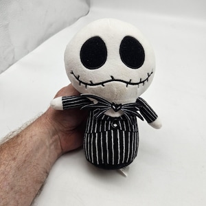 May include: A plush toy of Jack Skellington from The Nightmare Before Christmas. The toy has a white head with large black eyes and a stitched smile. It wears a black and white pinstripe suit with a bow tie.
