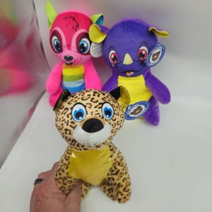 May include: Three plush animal toys: a pink deer with rainbow stripes, a purple rhino with yellow accents, and a leopard-print bear. Each toy has large, blue eyes and a tag.