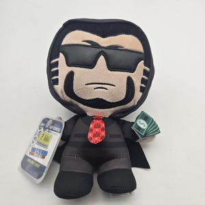 May include: A plush doll of a man wearing sunglasses, a black suit, and a red tie with a crown pattern. The doll holds a stack of green paper money in one hand and a tag that says "Jail Break" in the other.