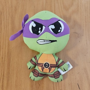 May include: A plush toy of a Teenage Mutant Ninja Turtle character, featuring a green head with a purple mask and a stern expression. The toy has an orange chest, brown accents, and the letter "D" on the belt. A small tag is attached.