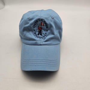 May include: Light blue baseball cap with a curved brim. The cap features embroidered text and a graphic of a golfer holding a flag. The text reads "124th U.S. OPEN PINEHURST".