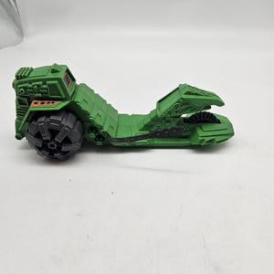 May include: A green toy vehicle with a large black wheel on the left side. The vehicle has a long, ridged body and a detailed front section. The toy is made of plastic and appears to be a vintage collectible.
