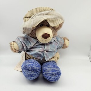 May include: A plush teddy bear wearing a tan hat with a white veil, a blue plaid shirt, khaki pants, and blue and white striped socks. The bear has a light brown nose and a tag attached to its clothing.