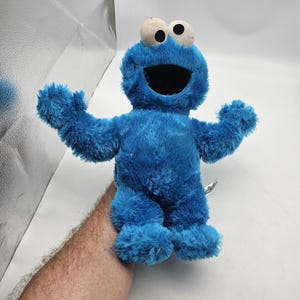 May include: A plush Cookie Monster toy in bright blue with large white eyes and a wide open black mouth. The toy has outstretched arms and fluffy blue fur. A classic Sesame Street character.