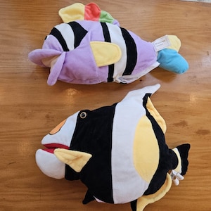 May include: Two plush fish toys. One is lavender with black stripes, yellow fins, and a rainbow tail. The other is black and white with yellow accents and a red mouth. Both are on a wooden surface.