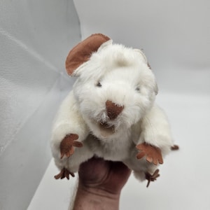 May include: A plush, white wombat stuffed animal with brown ears, nose, and paws. The toy has a soft, furry texture and is designed to resemble a real wombat. The stuffed animal is held in a hand.