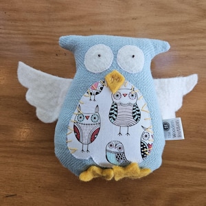 May include: A blue fabric owl-shaped plush toy with white felt wings and yellow feet. The owl has large white eyes and a decorative patch on its chest featuring various owl designs. The tag reads "The Wooden Button".