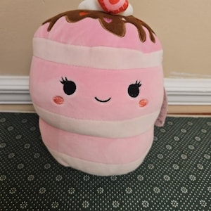 May include: A plush, pink cupcake-shaped pillow with white and pink stripes. It has a smiling face with black eyes and rosy cheeks. The top is decorated with brown frosting, white whipped cream, and a strawberry heart.