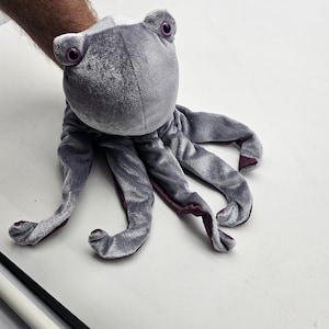 May include: A gray plush octopus puppet with large, purple eyes and eight tentacles. The puppet is made of soft, velvety material. The tips of the tentacles are a darker shade of purple.