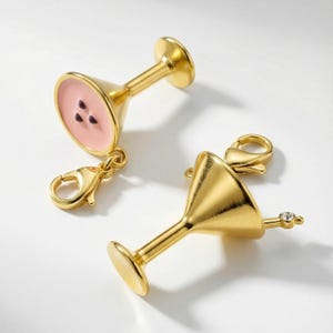 Könnte beinhalten: Zwei goldfarbene Martini-Anhänger. Einer hat eine rosa Innenseite mit drei dunkelbraunen Punkten, der andere einen klaren Edelstein am Stiel. Beide haben Karabinerverschlüsse für eine einfache Befestigung.