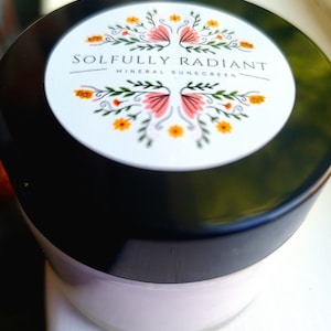 Puede incluir: Un tarro blanco con tapa negra y una etiqueta con diseño floral que dice "Soulfully Radiant Mineral Sunscreen".