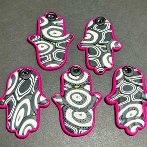 May include: Five Hamsa hand charms with a black, white, and gray swirl pattern, outlined in bright pink. Each charm has a small black button at the top. The charms are arranged on a gray surface.