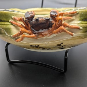 May include: A ceramic bowl with a crab design. The crab is orange and brown, with a dark brown body. The bowl has a green and tan background and is supported by a black metal stand. The bowl has Asian characters.